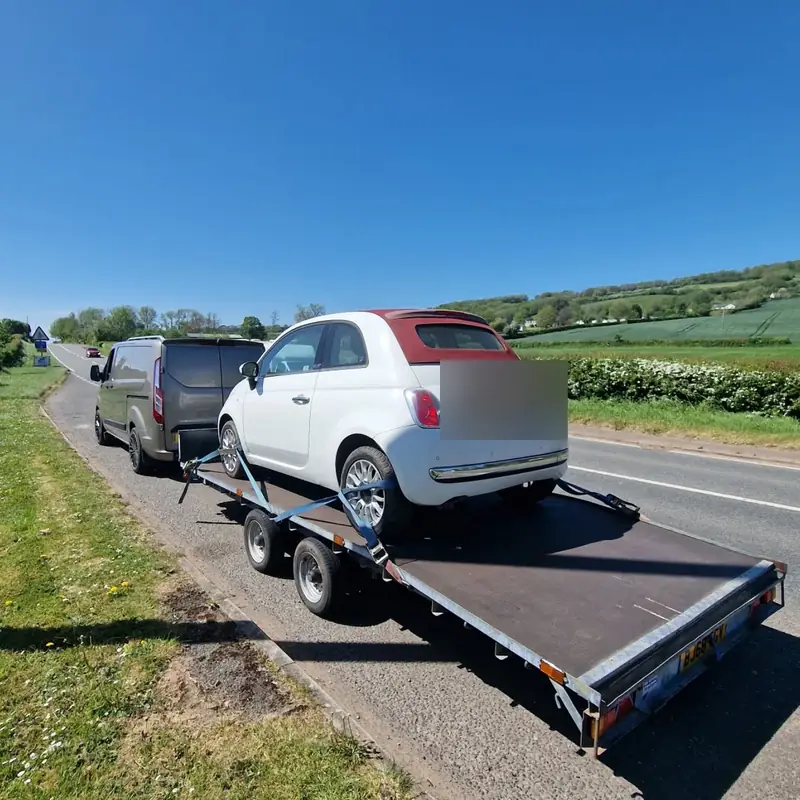 White Fiat 500 convertible strapped onto a flatbed trailer beside a country road
